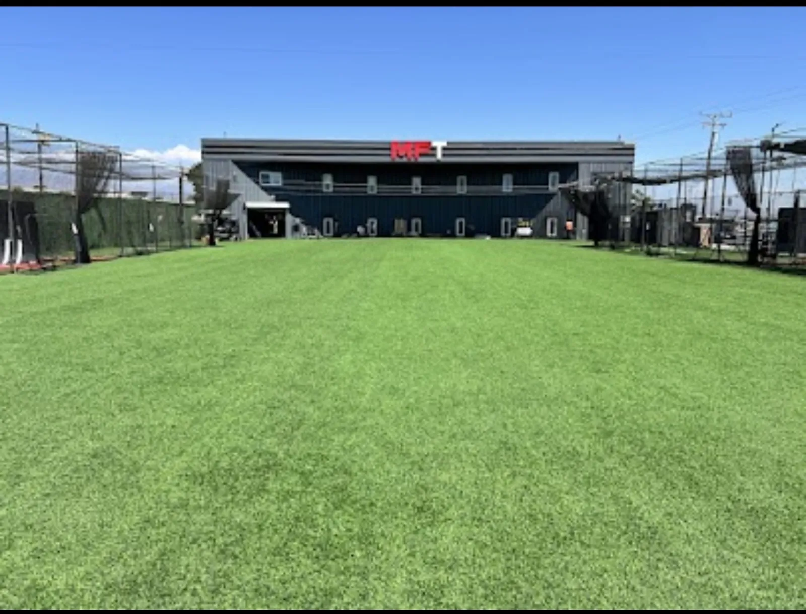 Outdoor turf training field at MF Training SB with batting cages along both sides and the MFT building behind